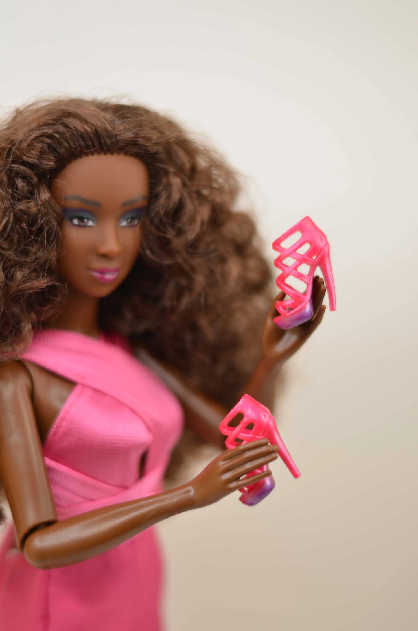 Barbie doll with brown hair and a pink dress holding pink high heels against a beige background