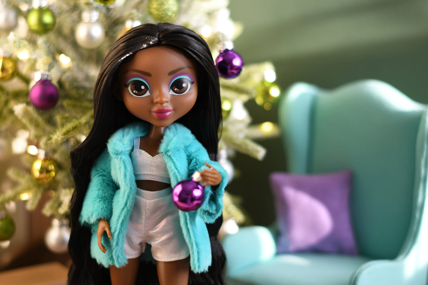 Doll wearing a turquoise coat in front of a decorated Christmas tree