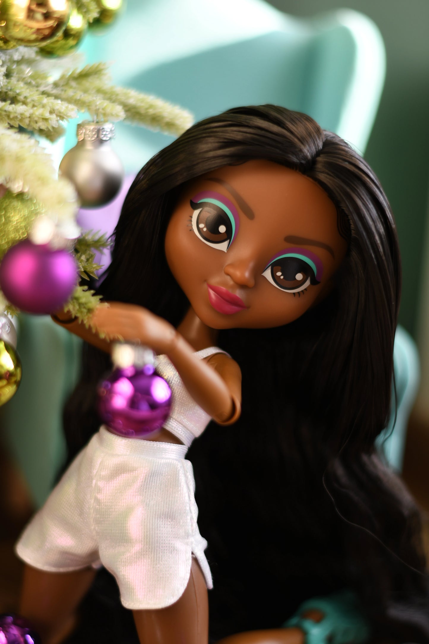 Doll with long dark hair holding a purple ornament in front of a decorated Christmas tree.