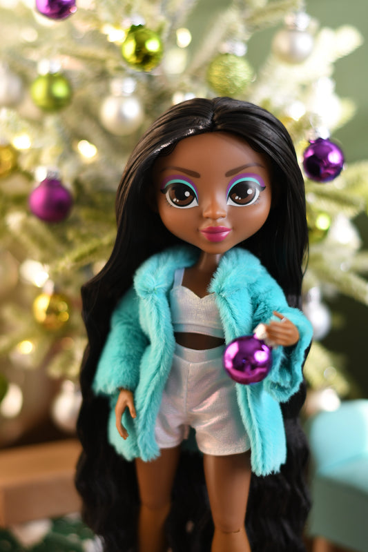 Doll wearing a turquoise coat in front of a decorated Christmas tree