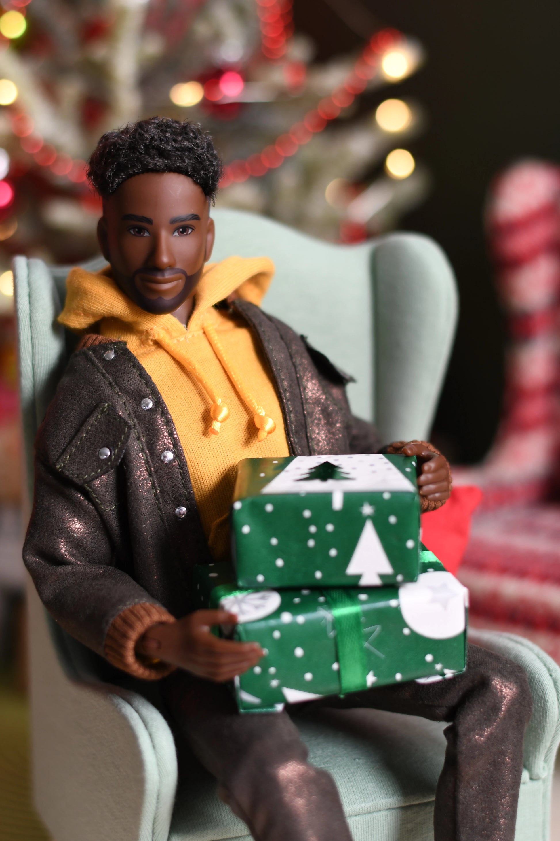 Action figure of a man in a brown coat and yellow sweater holding Christmas presents, with blurred festive background.